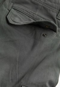 Dunkelgrauer Stoff mit glatter Textur, versehen mit einer Klapptasche, sichtbaren Nähten und einem metallischen Druckknopfdetail.