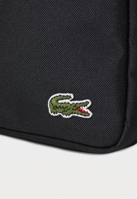 Svart teksturert stoffveske med en brodert grønn krokodillelogo med røde detaljer. Merkelig søm og strukturert form.