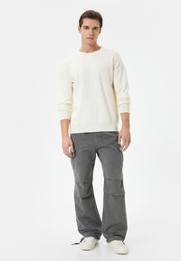 Suéter de punto color crema con puños acanalados, combinado con pantalones cargo grises que cuentan con bolsillos y un ajuste relajado. Zapatillas blancas completan el conjunto.