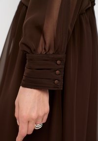 Bruine chiffon jurk met mouwen die een geraffineerde manchet hebben met drie knoopaccenten, een gladde textuur en een zacht, vloeiend materiaal. Hand met zilveren ring zichtbaar.