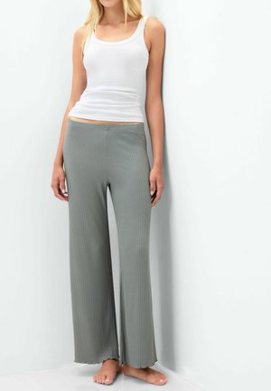 Vrouw draagt een witte mouwloze top en losse, geribbelde grijze broek, staat op blote voeten tegen een witte muur.