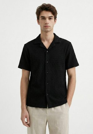 Ung mann iført en teksturert, kortermet svart skjorte med knapper og beige bukser, stående mot en enkel lys bakgrunn.