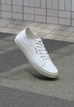 White canvas sneaker with round toe, lace-up front, and rubber sole. Features a small pull tab at the heel and subtle branding on the side.