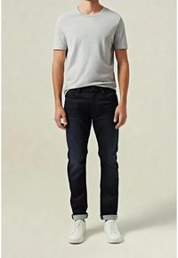 Mann trägt ein hellgraues T-Shirt mit kurzen Ärmeln, dunkel hochgekrempelte Jeans und weiße Turnschuhe, steht vor einem einfarbigen beigefarbenen Hintergrund.