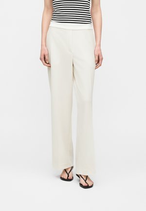 Woman wearing high-waist wide-leg cream trousers, black and white striped sleeveless top, and black thong sandals standing on white background.