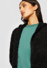 Veste à capuche noire en peluche avec une texture, superposée sur un haut vert uni. La veste n'a pas de fermetures visibles.