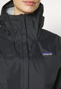 Giacca nera impermeabile con colletto alto, cappuccio regolabile e logo Patagonia. Caratterizzata da una texture liscia e lucida e da un design minimalista.