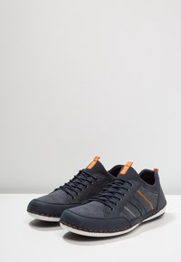 Ett par marinblå vardagssneakers med oranga dragflikar, orange sömmar, svarta snören och vita sulor på en vit yta.