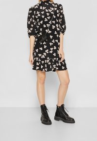 Černé květinové šaty s krátkými nabíranými rukávy, volánkovým lemem a béžovými vzory. V páru s černými šněrovacími kotníkovými botami. Malá černá crossbody kabelka.