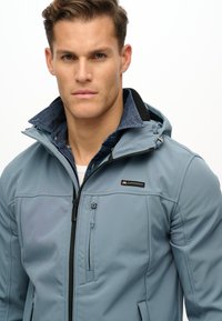 Veste à capuche bleu clair avec fermeture éclair frontale, présentant un col noir, un logo sur la poitrine et plusieurs poches. Fabriquée en tissu lisse et durable.