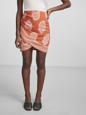 Orangenes Wickelrock mit weißem Paisley-Muster und horizontalem Streifen am Saum. Glattes Material mit einer figurbetonten Silhouette. Grüne Riegelsandalen.