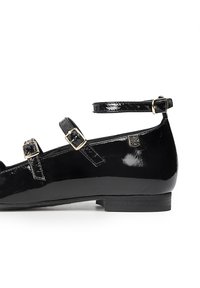 Zapato plano de charol negro con tira al tobillo, tres hebillas ajustables, punta redondeada y tacón bajo. Presenta un pequeño detalle de logo.