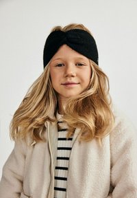 Fascia in maglia nera con dettaglio annodato sul davanti, indossata da un bambino. La texture morbida contrasta con un leggero giubbotto in pile sopra una maglia a righe.