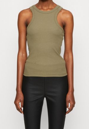 Femme portant un haut côtelé sans manches vert olive et un pantalon en cuir noir taille haute, debout devant un fond clair uni.