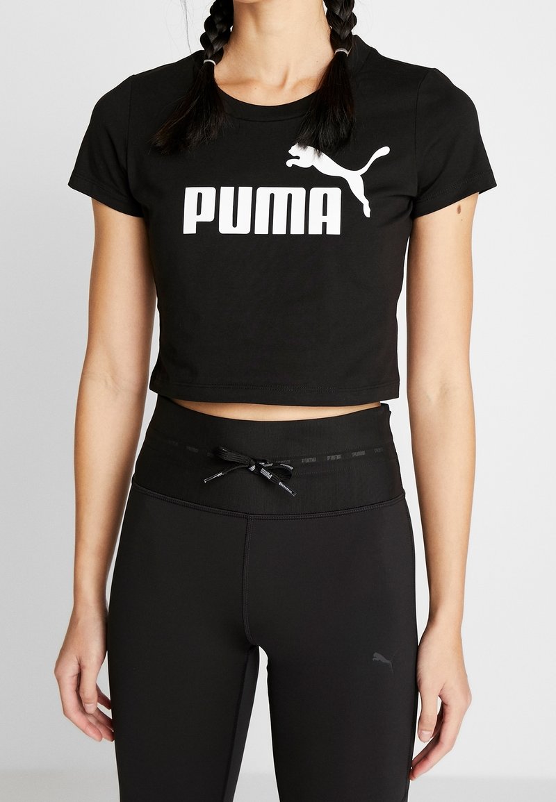 Jeune femme portant un t-shirt court Puma noir et un legging noir taille haute avec cordon à la taille, sur fond blanc.