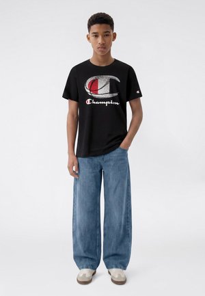 Junge Person steht und trägt ein schwarzes Champion-Logo-T-Shirt, weite blaue Jeans und beige Sneakers vor einem einfarbigen weißen Hintergrund.
