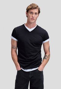 Jaunas vyras su šviesiai ruda plaukais, vilkintis sluoksniuotus juodus ir baltus marškinėlius, juodus džinsus, su tatuiruotėmis ant abiejų dilbių, rankos kišenėse.