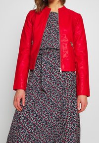 Veste en cuir rouge avec fermeture éclair sur le devant, présentant des manches longues et un design ajusté. Portée par-dessus une robe bleu marine à imprime floral avec une taille à nouer.