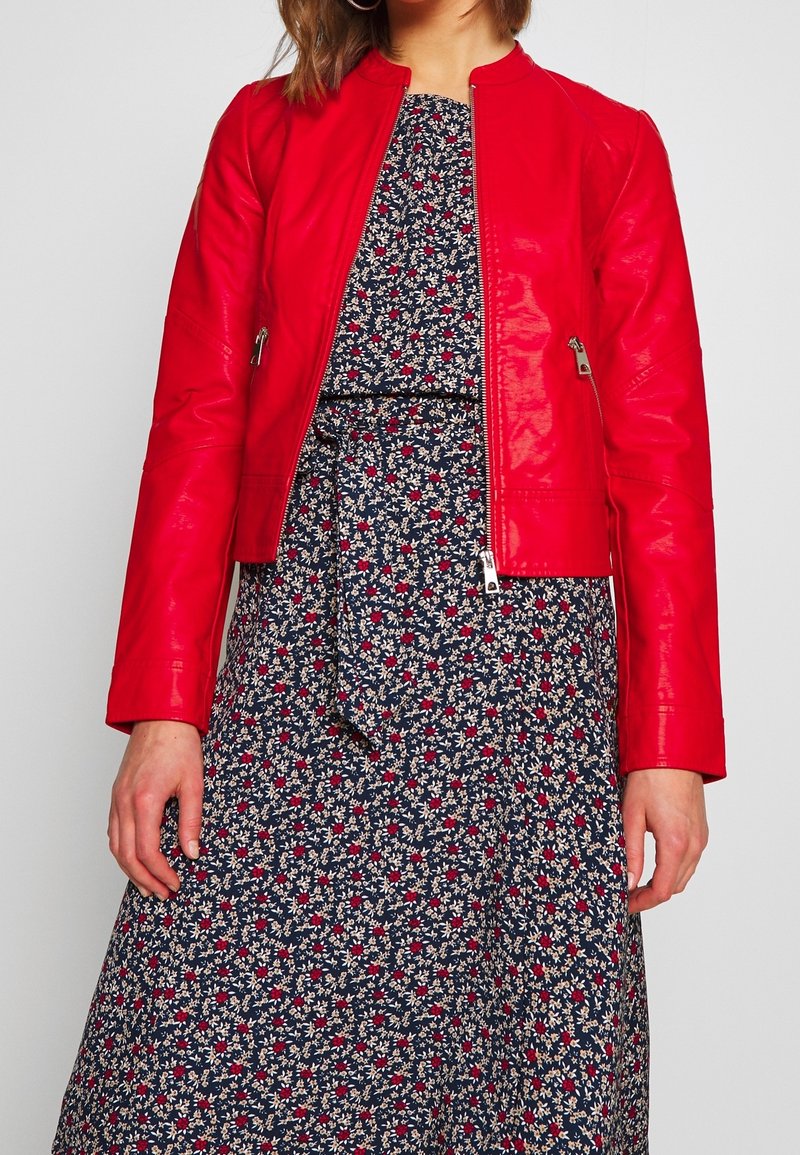 Veste en cuir rouge avec fermeture éclair sur le devant, présentant des manches longues et un design ajusté. Portée par-dessus une robe bleu marine à imprime floral avec une taille à nouer.