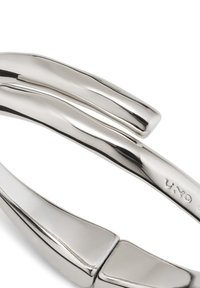 Pulsera de metal plateado con un diseño ondulado y suave. Presenta dos segmentos curvados unidos por una bisagra, reflejando la luz con un acabado pulido.