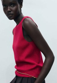 Aangesloten rode gebreide tanktop met ruime armsgaten, voorzien van een gouden kettingaccent in de taille. Glad textuur, minimalistisch ontwerp.