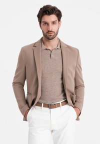 Beiger Blazer über einem hellbraunen Polohemd, weißen Hosen und einem braunen Gürtel. Glatte Stoffqualität mit taillierter Passform und subtilen Details.