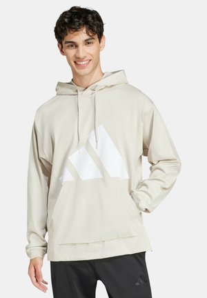 Giovane uomo sorridente, indossa una felpa con cappuccio beige con un grande logo bianco e pantaloni neri con un piccolo logo, in piedi davanti a uno sfondo neutro.