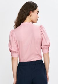 Różowa bluzka z bufkami na krótkich rękawach, gładka w dotyku, z ozdobnymi guzikami i kołnierzykiem. Połączona z ciemnogranatowymi spodniami, prezentowana z tyłu.