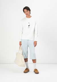 Jonge man in een witte lange mouw shirt met een zwart-wit foto afdruk, lichte denim shorts, bruine schoenen en een beige tote bag in de hand.