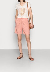 T-shirt blanc à col en V avec un motif de cœur. Shorts en lin rose avec une taille à cordon. Sandales brunes à lanières. Petit sac bandoulière marron.