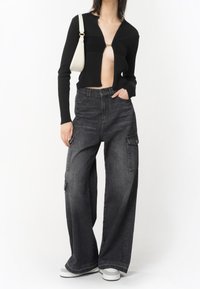 Cardigan noir à côtes avec un décolleté en V profond et des ornements en or, associé à un pantalon cargo en denim gris ample avec poches latérales et une coupe décontractée.