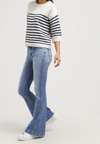 Kvinna som bär en vit och marinblå randig tröja, ljusblå utsvängda jeans och vita sneakers, stående mot en enfärgad bakgrund.