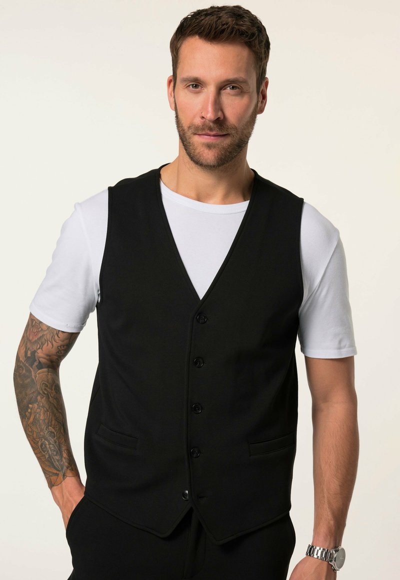 Chaleco negro con diseño de escote en V, cinco botones y dos bolsillos laterales. Lleva encima una camiseta blanca. Presenta una textura de tela suave.