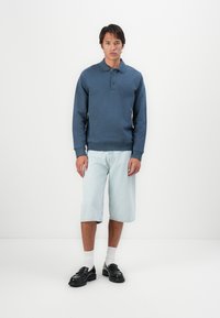 Blå hettegenser med krage og to knapper, sammen med lyseblå knelange denimshorts og svarte kraftige sko. Hvite sokker synlige.