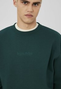 Sweat-shirt vert foncé avec un col rond et des poignets côtelés. Présente un logo en relief, "légendaire," sur la poitrine. Texture lisse.