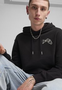 Felpa nera in cotone con cappuccio, lacci metallici e grafica ricamata che presenta un pallone da basket e dettagli. Abbinata a jeans azzurri chiari.