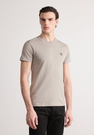 Beige Baumwoll-T-Shirt mit kurzen Ärmeln, Rundhalsausschnitt und kleinem schwarzen Logo auf der linken Brust; glatte Textur, taillierte Passform.