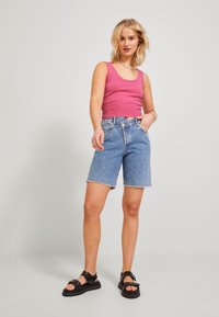 Top a righe rosa, pantaloni shorts in denim blu con orlo sfrangiato e sandali neri spessi. Modella in piedi davanti a uno sfondo neutro.