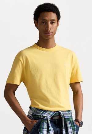 Gul bomulds T-shirt med rund hals, der har korte ærmer. Modellen har en ternet skjorte bundet om livet og slidte jeans på.