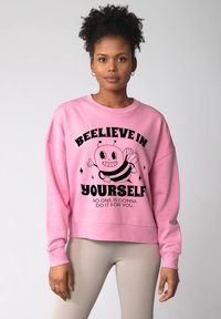Roze sweatshirt met een zwart cartoonbijtje en de tekst "GELÓÓF IN JEZELF" erboven. Voorzien van geribbelde manchetten en een ontspannen pasvorm.
