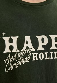 T-shirt à manches courtes vert foncé avec un texte blanc indiquant "JOYEUSES FÊTES" et "Et joyeux Noël" dans diverses polices. Texture douce.