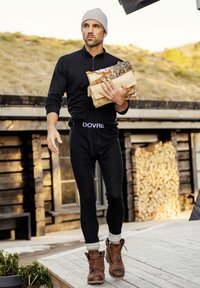 Schwarzes Reißverschluss-Hemd mit langen Ärmeln und schwarze Leggings mit einem Logo-Bund, braune Stiefel, graue Mütze, trägt Feuerholz im Freien.