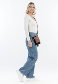 Borsa a tracolla nera e marrone con dettagli di zip; il modello indossa una maglietta bianca aderente e jeans a gamba larga blu chiaro, abbinati a sneakers bianche.