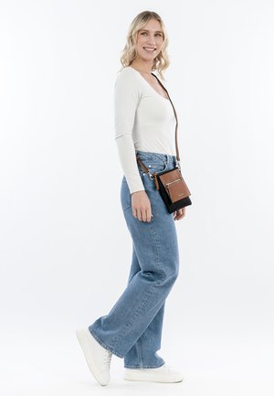 Black and brown crossbody bag with zipper accents; model wears a fitted white top and light blue wide-leg jeans, paired with white sneakers.