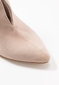 Béžová semišová kotníková obuv s špičatou špičkou, s výřezovým designem na straně a viditelným švem podél švů.