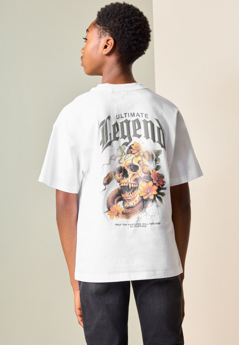 Junge Person trägt ein weißes T-Shirt mit Totenkopf, Schlange, orangefarbenen Blumen und dem Text "Ultimate Legend" auf dem Rücken.