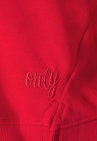 Tela de algodón roja con textura acanalada y detalle de costura. La palabra "only" está bordada con un hilo rojo oscuro contrastante.