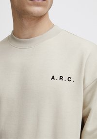 Zbliżenie na osobę noszącą beżowy sweter z czarnym napisem "A.R.C." na lewej piersi oraz widoczną srebrną łańcuszek na szyi.