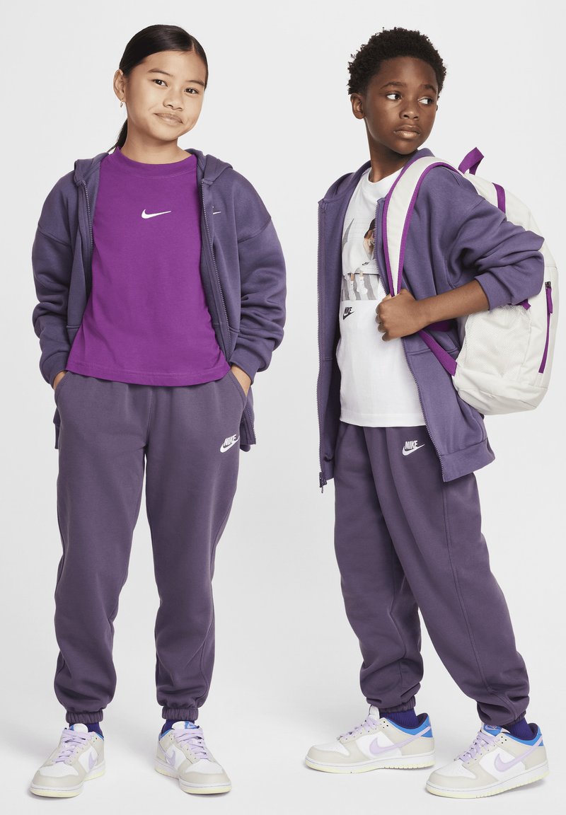 Két gyermeken lila Nike kapucnis felsők és melegítők; az egyikük fehér grafikás pólót és krém színű hátizsákot visel, lila részletekkel.