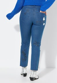Blauwe spijkerbroek met een slim fit, voorzien van witte hartpatches en zwarte bedrukte zinnen. Inclusief een rafelrand en twee achterzakken.